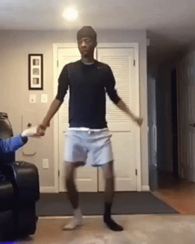 A Man In A Black Shirt And Blue Shorts Is Dancing In A Living Room . GIF