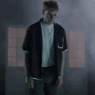 A Man In A Black Shirt And Glasses Is Standing In A Dark Room . GIF