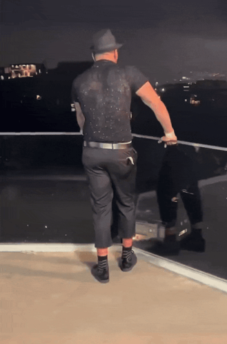 A Man In A Black Shirt And Hat Is Standing On A Ledge GIF