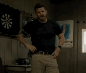 A Man In A Black Shirt And Khaki Pants Is Standing In Front Of A Dart Board In A Room . GIF
