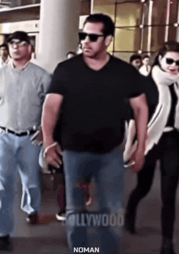 A Man In A Black Shirt And Sunglasses Is Walking With A Woman In A White Scarf Behind Him . GIF