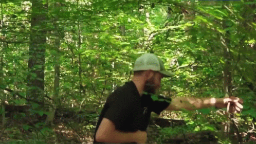 a man in a black shirt and white hat is walking through the woods .