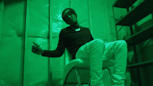 A Man In A Black Shirt And White Pants Is Sitting In A Green Room GIF