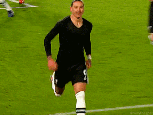 A Man In A Black Shirt And White Socks Stands On A Soccer Field Next To A Red Flag GIF