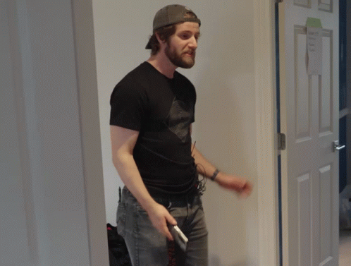 A Man In A Black Shirt Holds A Cell Phone In Front Of A Door GIF