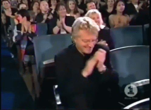A Man In A Black Shirt Is Clapping In Front Of A Crowd With A Number 1 On Their Seat GIF