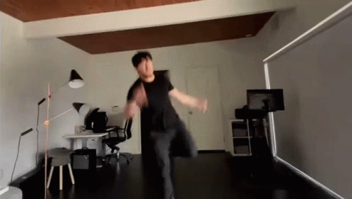 A Man In A Black Shirt Is Dancing In A Room With A Desk And Chair GIF