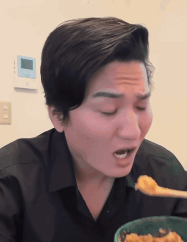 A Man In A Black Shirt Is Eating A Bowl Of Food With A Spoon GIF