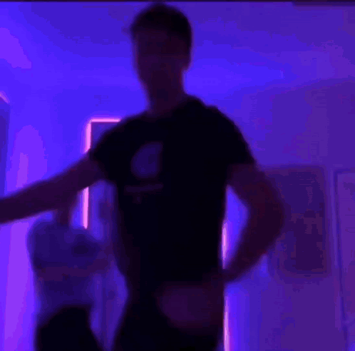 A Man In A Black Shirt Is Flexing His Muscles In A Room With Purple Lights . GIF