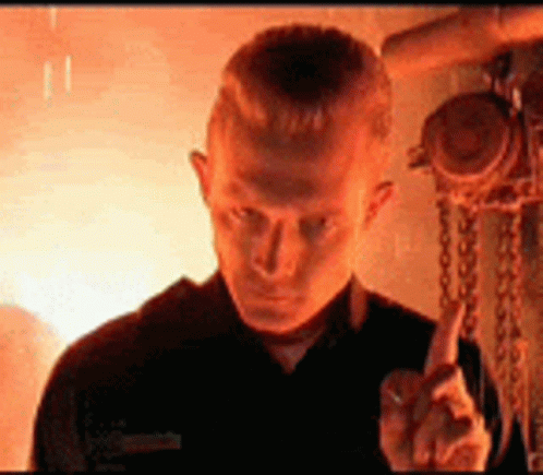 A Man In A Black Shirt Is Giving The Middle Finger In Front Of A Fire . GIF