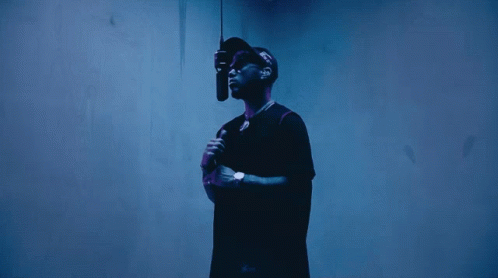 A Man In A Black Shirt Is Holding A Microphone In Front Of A Blue Wall GIF
