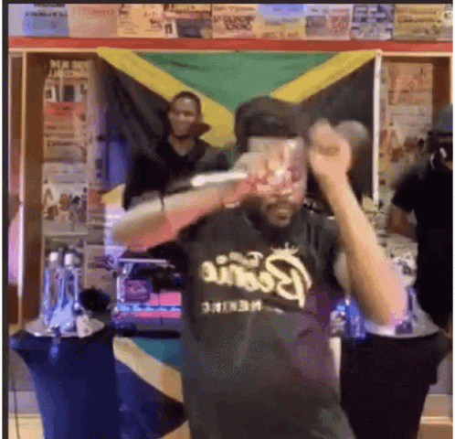 A Man In A Black Shirt Is Holding A Microphone In Front Of A Flag . GIF
