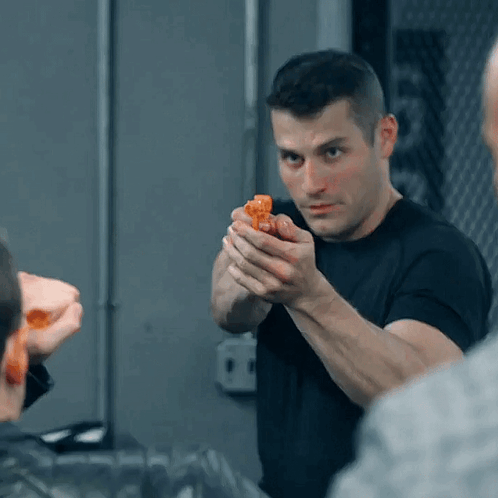 A Man In A Black Shirt Is Holding A Small Orange Object In His Hand GIF