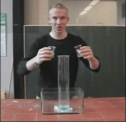 A Man In A Black Shirt Is Holding Two Cups In Front Of A Glass Container GIF