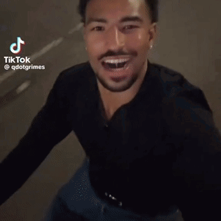 A Man In A Black Shirt Is Laughing And Smiling While Standing On A Street . GIF