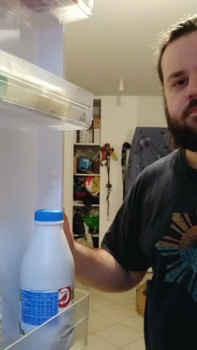 A Man In A Black Shirt Is Reaching Into A Fridge To Take A Bottle Of Milk GIF