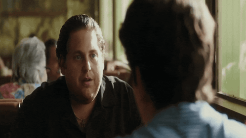 a man in a black shirt is sitting at a table talking to another man
