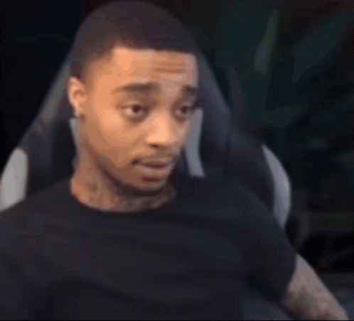 A Man In A Black Shirt Is Sitting In A Chair And Looking At The Camera . GIF
