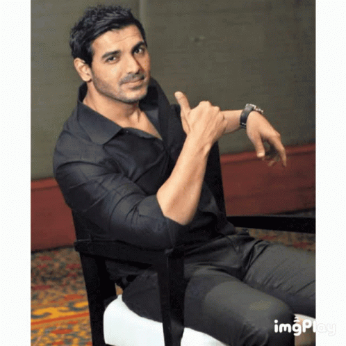 A Man In A Black Shirt Is Sitting In A Chair And Pointing At His Wrist GIF
