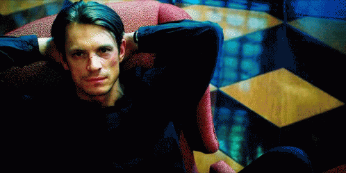 A Man In A Black Shirt Is Sitting In A Chair With His Hands Behind His Head GIF