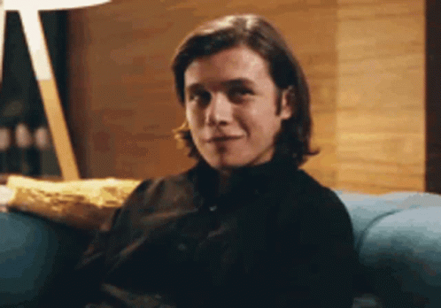 A Man In A Black Shirt Is Sitting On A Blue Couch Looking At The Camera . GIF
