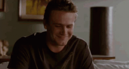 A Man In A Black Shirt Is Sitting On A Couch And Smiling . GIF
