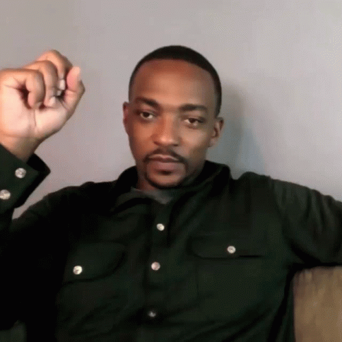 A Man In A Black Shirt Is Sitting On A Couch With His Fist Up GIF