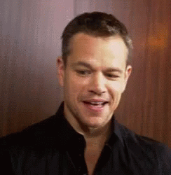 A Man In A Black Shirt Is Smiling And Looking At The Camera While Standing In Front Of A Wooden Wall . GIF