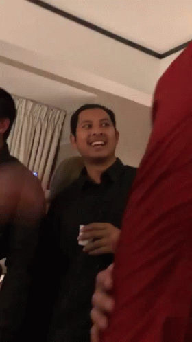 A Man In A Black Shirt Is Smiling In A Room With Other People GIF
