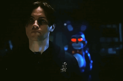 A Man In A Black Shirt Is Smiling In Front Of A Stuffed Animal That Has Red Eyes GIF