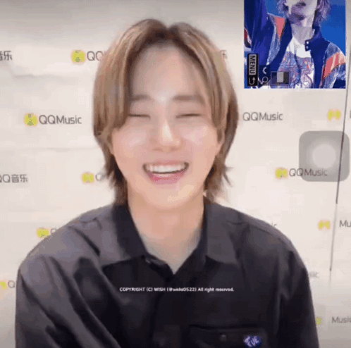 A Man In A Black Shirt Is Smiling In Front Of A Wall That Says Qqmusic GIF