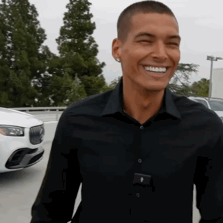 A Man In A Black Shirt Is Smiling In Front Of A White Car In A Parking Lot GIF