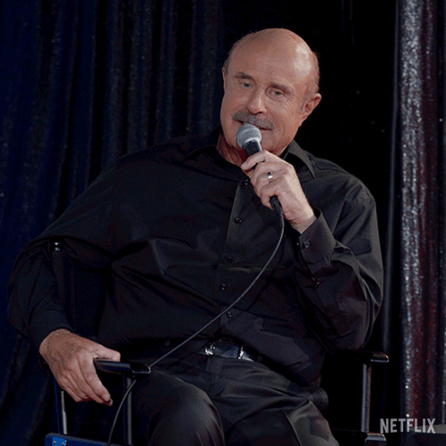 A Man In A Black Shirt Is Speaking Into A Microphone With A Netflix Logo In The Background GIF