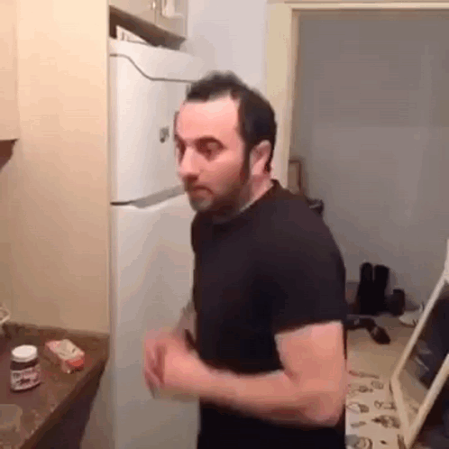 A Man In A Black Shirt Is Standing In A Kitchen Next To A Refrigerator . GIF
