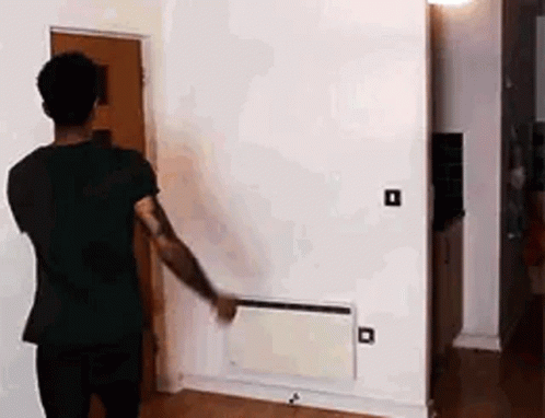 A Man In A Black Shirt Is Standing In A Living Room Looking At A Wall Heater . GIF