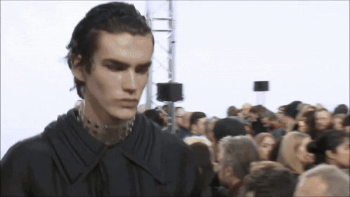 A Man In A Black Shirt Is Standing In Front Of A Crowd Of People GIF
