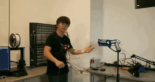 A Man In A Black Shirt Is Standing In Front Of A Desk With A Computer And A Gun On It GIF