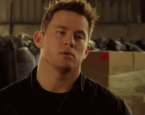 A Man In A Black Shirt Is Standing In Front Of A Pile Of Boxes And Looking At The Camera . GIF