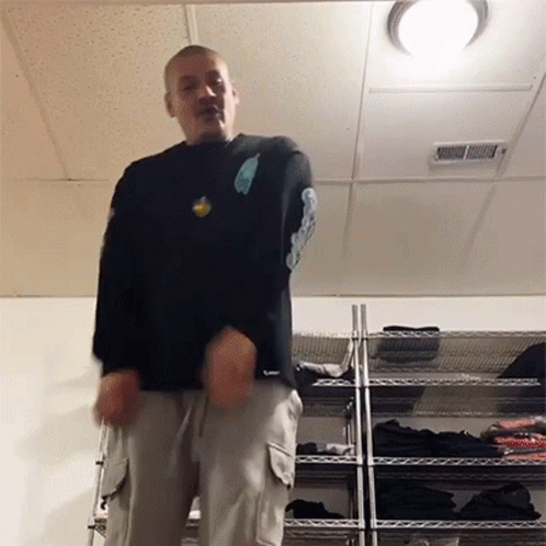 A Man In A Black Shirt Is Standing In Front Of A Rack Of Clothes GIF