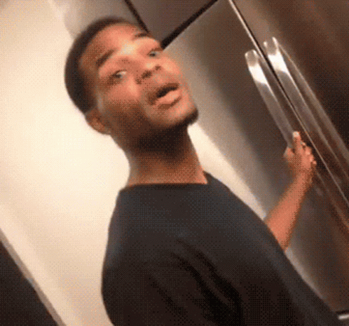 A Man In A Black Shirt Is Standing In Front Of A Refrigerator With His Mouth Open GIF