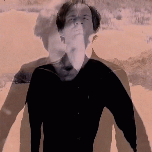 A Man In A Black Shirt Is Standing In Front Of A Sandy Beach GIF