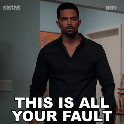 A Man In A Black Shirt Is Standing In Front Of A Sign That Says This Is All Your Fault GIF