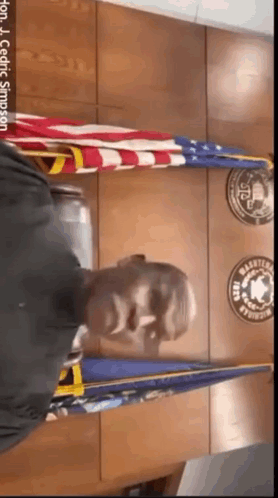 A Man In A Black Shirt Is Standing In Front Of A Wall With Flags And Badges On It .. GIF