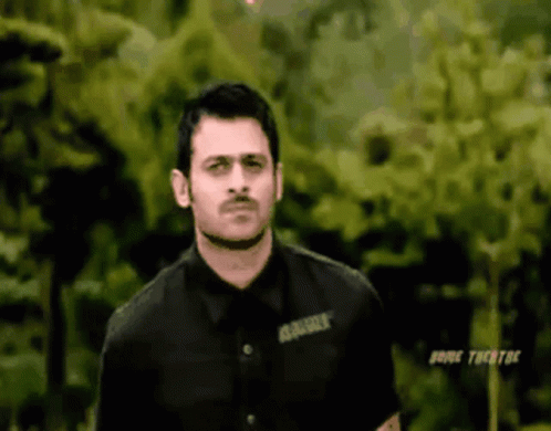 A Man In A Black Shirt Is Standing In Front Of Some Trees . GIF