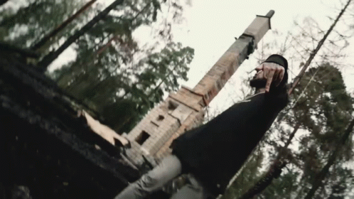 A Man In A Black Shirt Is Standing In The Woods In Front Of A Statue . GIF