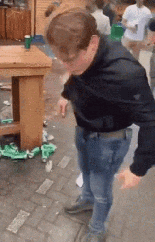 A Man In A Black Shirt Is Standing On A Brick Sidewalk In Front Of A Wooden Table . GIF