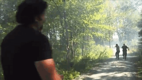 A Man In A Black Shirt Is Standing On A Dirt Road In The Woods Looking At Two Men Walking Down The Road . GIF