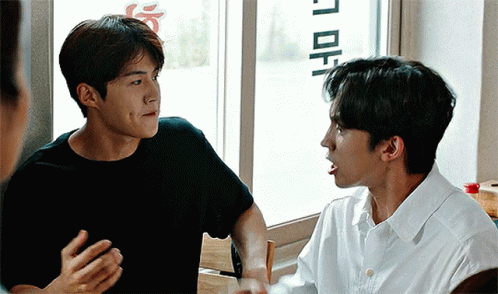 A Man In A Black Shirt Is Talking To Another Man In A White Shirt While Sitting In Front Of A Window . GIF