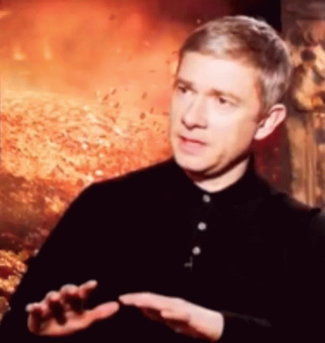 A Man In A Black Shirt Is Talking To Someone In Front Of A Fire Background . GIF