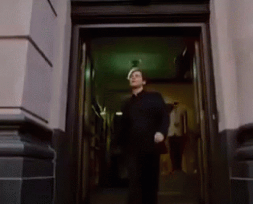 A Man In A Black Shirt Is Walking Out Of A Doorway . GIF